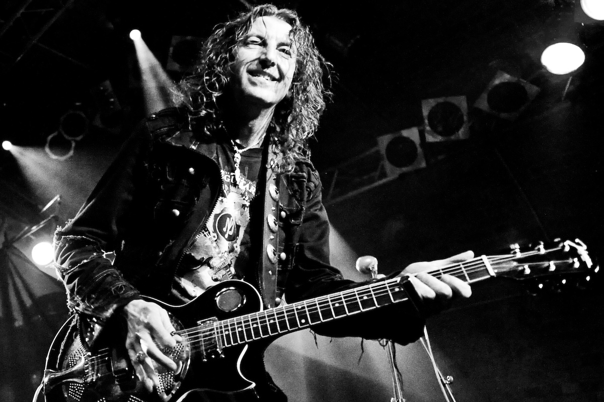 Blues rock guitarist Gwyn Ashton smiling on stage as he plays a resonator guitar under dramatic concert lights.